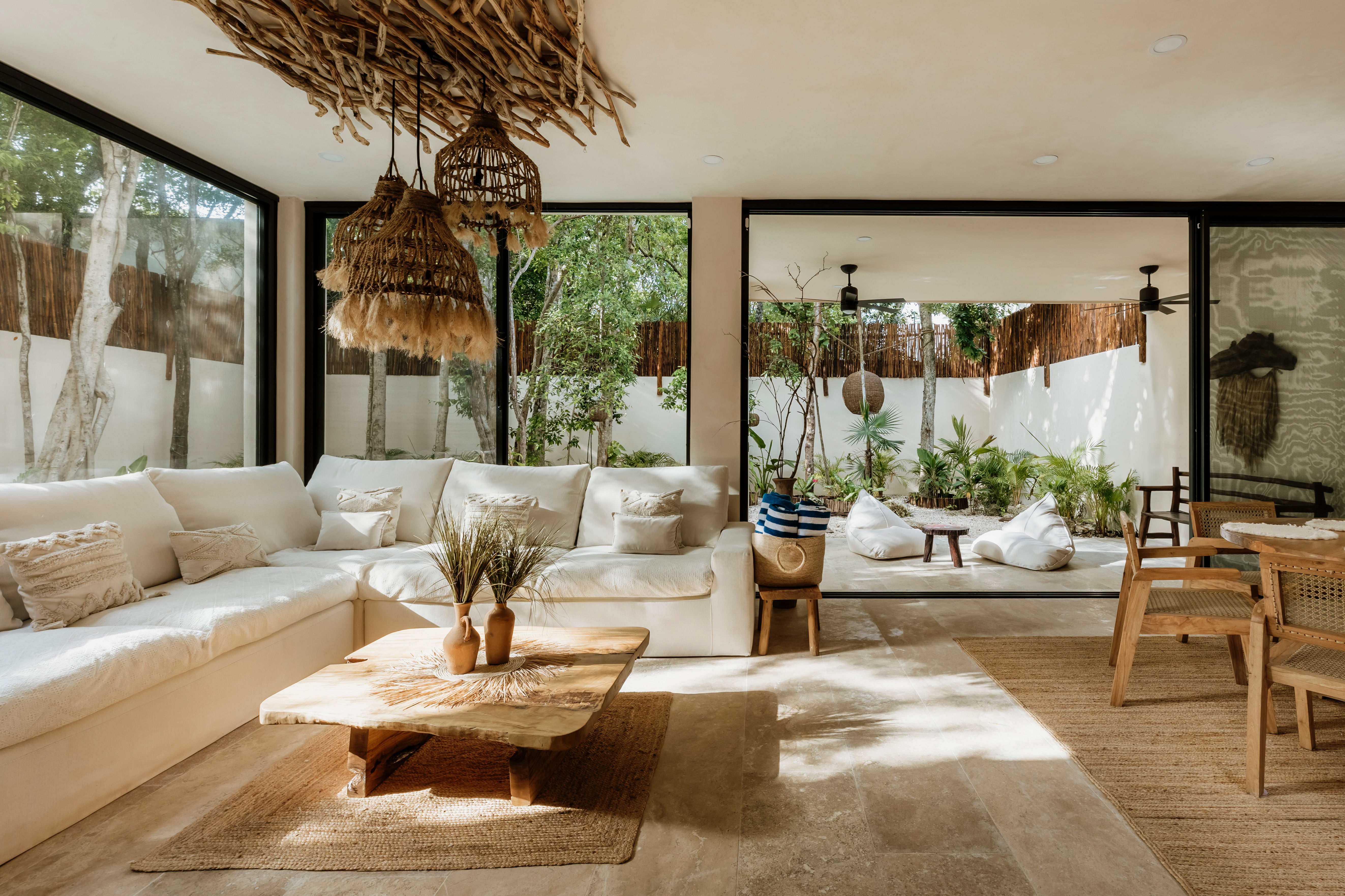 Sunlit modern boho living room with white sectional sofa, driftwood rattan pendant lights, wooden coffee table and sliding glass doors opening to a lush patio with tropical plants and beanbags.
