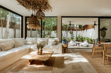 Sunlit modern boho living room with white sectional sofa, driftwood rattan pendant lights, wooden coffee table and sliding glass doors opening to a lush patio with tropical plants and beanbags.