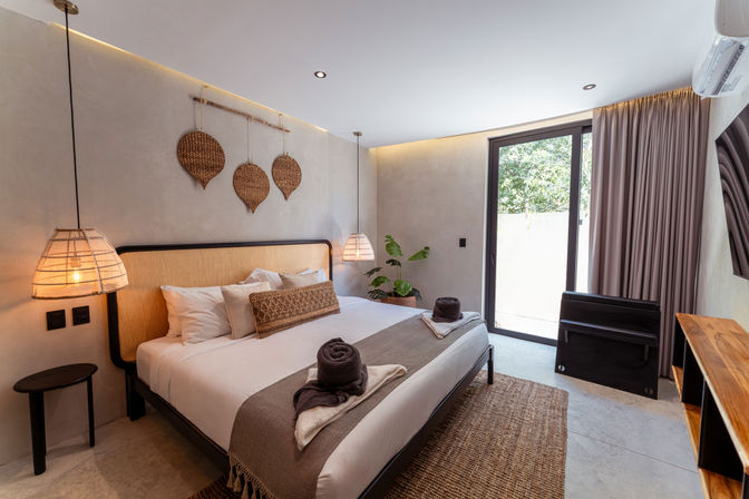 Cozy modern bedroom with king-size bed, woven headboard and wicker wall hangings, rattan pendant lamps, neutral linens and textured rug, potted plant and sliding glass door to leafy outdoor views.
