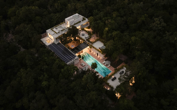 Aerial dusk view of a modern luxury villa and rooftop terraces with solar panels, glowing turquoise swimming pool and outdoor lounge areas tucked into a secluded, lush forest retreat.