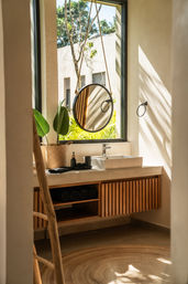 Sunlit modern bathroom vanity in a tropical-style villa — hanging round rope mirror, white vessel sink, woven plant basket, wooden slatted cabinet and rolled towels.