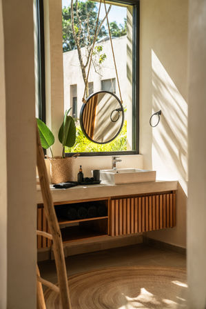 Sunlit modern bathroom vanity in a tropical-style villa — hanging round rope mirror, white vessel sink, woven plant basket, wooden slatted cabinet and rolled towels.