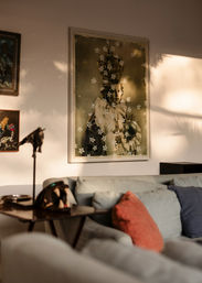 Sunlit living room with a light-gray sectional, coral and navy throw pillows, wooden side table with lamp and decorative object, and a large framed vintage portrait of a crowned child holding an orb overlaid with small white flowers.