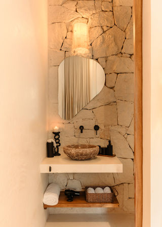 Mediterranean-style powder room with rough stone wall, organic stone vessel sink on a floating plaster shelf, sculptural irregular mirror, matte black fixtures, warm ambient lighting, candle and wicker towel basket.