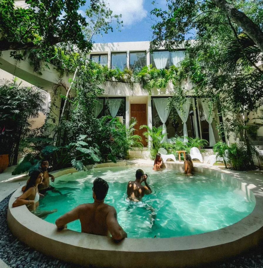 Sunlit circular courtyard pool at a tropical villa surrounded by lush greenery, floor-to-ceiling windows with white curtains, and people relaxing in the water — a peaceful outdoor urban oasis.