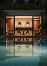 Nighttime pool reflection of a modern tropical villa’s warm-lit indoor lounge with round mirrors above a console, paired mid-century chairs and a small table, framed by lush plants.