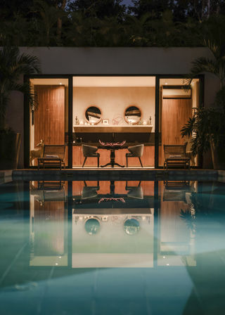 Nighttime pool reflection of a modern tropical villa’s warm-lit indoor lounge with round mirrors above a console, paired mid-century chairs and a small table, framed by lush plants.