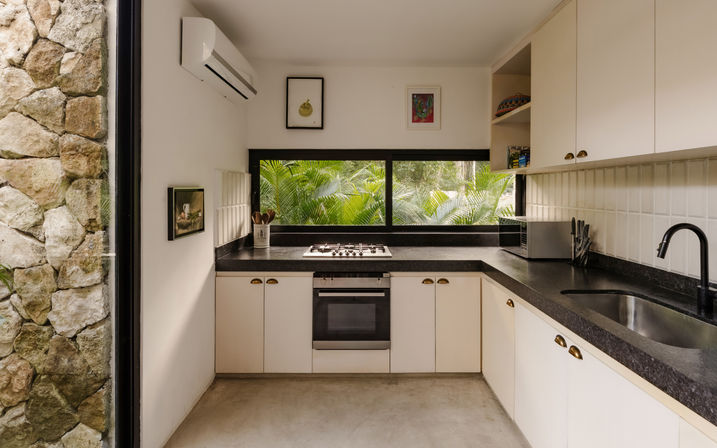 Minimal modern kitchen with black countertops and white cabinets, built-in oven and gas cooktop, long horizontal window framing lush tropical palm foliage and a textured stone wall.