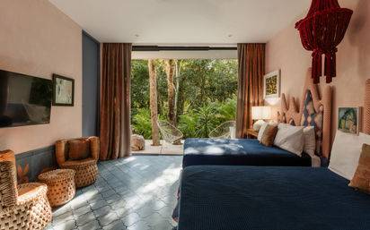 Tropical hotel room with two navy-blue beds, coral patterned headboards, woven rattan chairs and ottomans, blue tile floor and pink walls, opening onto a terrace with white outdoor chairs and lush green forest view.