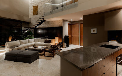 Luxury modern open-plan living room and kitchen with an L-shaped beige sectional, dark ottoman, wood coffee table, floating metal staircase on a stone accent wall, and a marble-topped island under warm ambient lighting.
