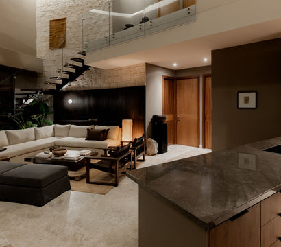 Luxury modern open-plan living room and kitchen with an L-shaped beige sectional, dark ottoman, wood coffee table, floating metal staircase on a stone accent wall, and a marble-topped island under warm ambient lighting.