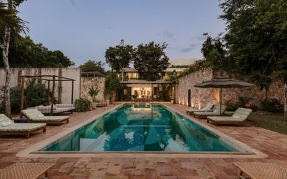 Inviting rectangular swimming pool at a tropical villa at dusk, flanked by stone walls, cushioned loungers and umbrellas, lush trees and warm interior lights glowing inside the house.