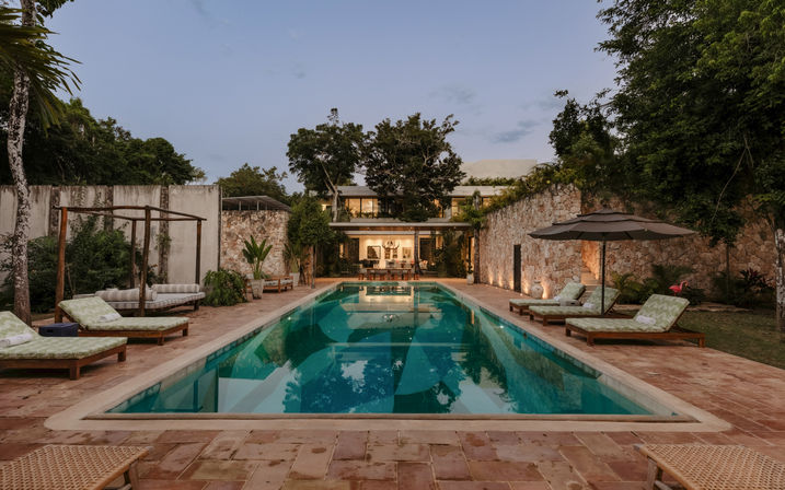 Inviting rectangular swimming pool at a tropical villa at dusk, flanked by stone walls, cushioned loungers and umbrellas, lush trees and warm interior lights glowing inside the house.