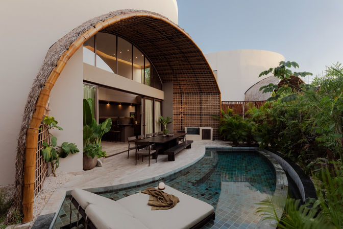 Modern tropical villa courtyard with a curved bamboo-and-thatch arch shading a wooden outdoor dining table, a cushioned lounge bed beside a tiled private plunge pool, lush tropical plants and sliding glass doors to the kitchen.