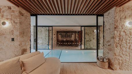 Indoor pool room with natural stone walls, wooden slatted ceiling and sliding glass doors opening to a long dining area; cozy beige sofa and woven towel basket in a Mediterranean-style luxury interior.