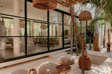 Sunlit tropical courtyard patio with floor-to-ceiling glass wall revealing an indoor dining area, woven rattan pendant lights, wooden stools and round beige floor cushions in a boho-modern lounge.