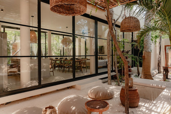 Sunlit tropical courtyard patio with floor-to-ceiling glass wall revealing an indoor dining area, woven rattan pendant lights, wooden stools and round beige floor cushions in a boho-modern lounge.