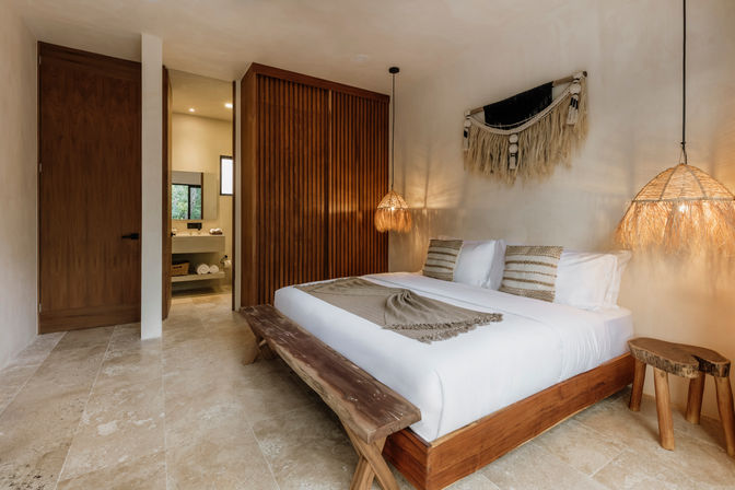 Modern boho bedroom with a wooden platform bed and white linens, striped throw pillows, woven wall hanging, rattan pendant lights, stone tile floor, rustic bench and open doorway to an en-suite bathroom.