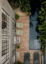 Top-down view of a narrow wooden poolside deck at a modern concrete villa with two lounge chairs, two green beanbag seats, potted plants and a reflective plunge pool edged by tropical foliage