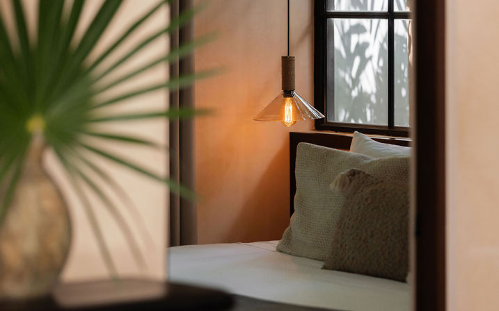 Cozy bedroom nook with a glowing pendant light, textured neutral pillows on a white bed, soft window shadows and a blurred palm leaf in the foreground.