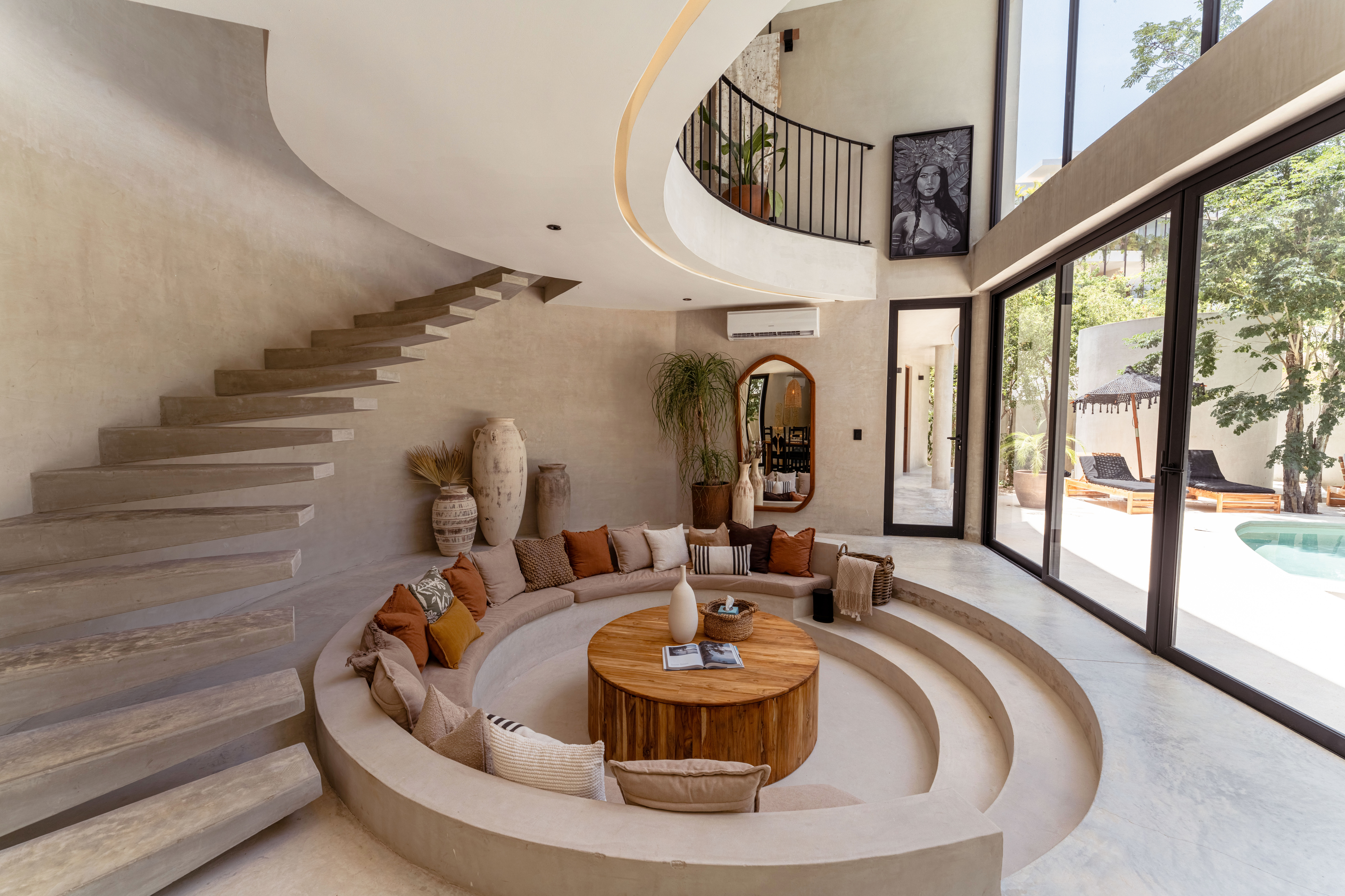 Sunken circular living room in a modern coastal villa with curved concrete tiers, built-in sofa and earth-tone pillows, floating staircase and floor-to-ceiling glass doors opening to a poolside patio with loungers.