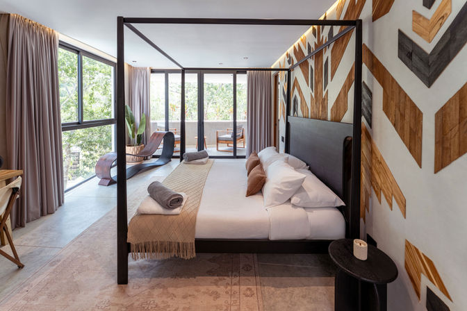 Sunlit contemporary bedroom with black four-poster bed, beige linens and rolled towels, geometric wood accent wall, floor-to-ceiling windows and balcony seating overlooking lush greenery.