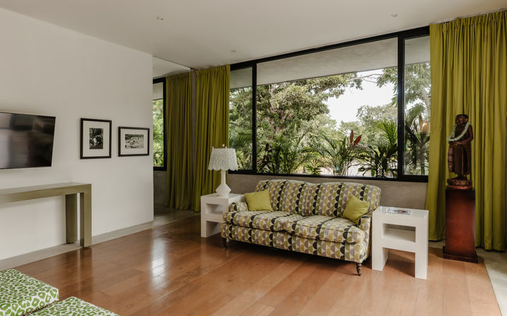 Sunlit contemporary living room with patterned green-gray sofa, floor-to-ceiling windows framing tropical garden, olive green curtains, white side tables, lamp and hardwood floor.