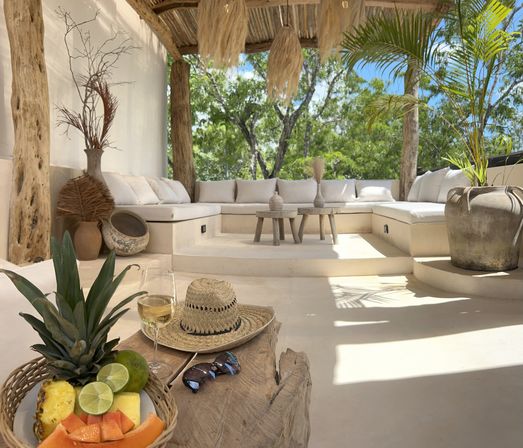 Sunlit tropical patio lounge with built-in white cushioned seating under a thatched wooden pergola, potted palms and rustic tables; foreground wooden table with fresh tropical fruit, a glass of white wine, straw hat and sunglasses.