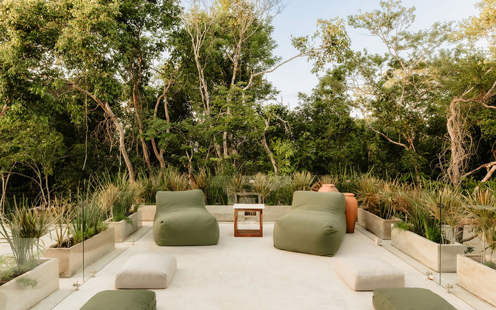 Modern outdoor terrace with olive-green beanbag lounge chairs and neutral floor cushions around a small wooden side table, flanked by concrete planters and lush trees for a relaxed patio vibe.
