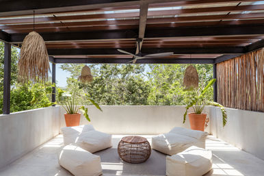 Sunlit tropical outdoor lounge with wood-beamed pergola, rattan pendant lights and ceiling fan, white beanbag cushions around a wicker coffee table and terracotta potted plants overlooking lush green trees.