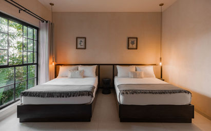 Minimalist guestroom with two identical beds dressed in white linens and gray throws, warm hanging pendant lights, framed botanical prints, and a large black-framed window overlooking lush green foliage.