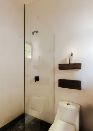 Minimalist bathroom with frameless glass shower, matte black shower fixtures, white one-piece toilet, rustic floating wooden shelves holding soap dispensers, and soft natural light on neutral plaster walls.