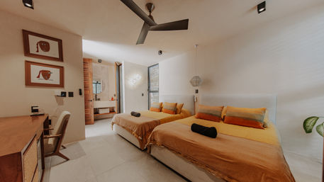 Sunlit modern twin hotel room with warm orange bedding, black ceiling fan, wooden desk, pendant light, en-suite bathroom with round mirror, and tropical potted plant.