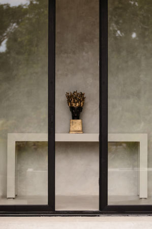 Minimalist storefront window display featuring a black bust sculpture topped with an ornate gold crown on a white console table against a textured concrete wall, modern interior styling.