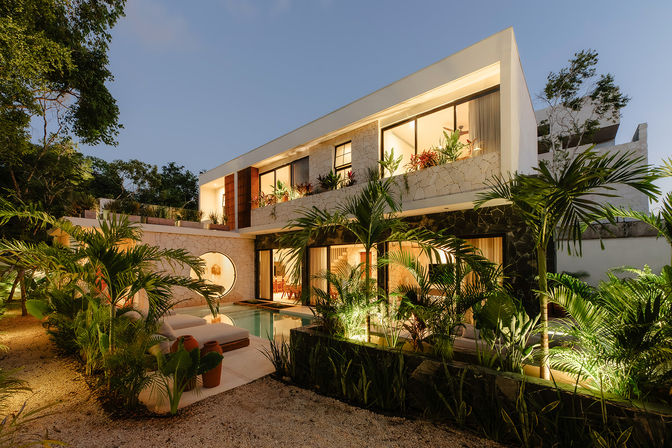 Modern two-story tropical villa at dusk with warm interior lighting, stone facade, floor-to-ceiling windows, private pool, outdoor lounge and lush palm landscaping.