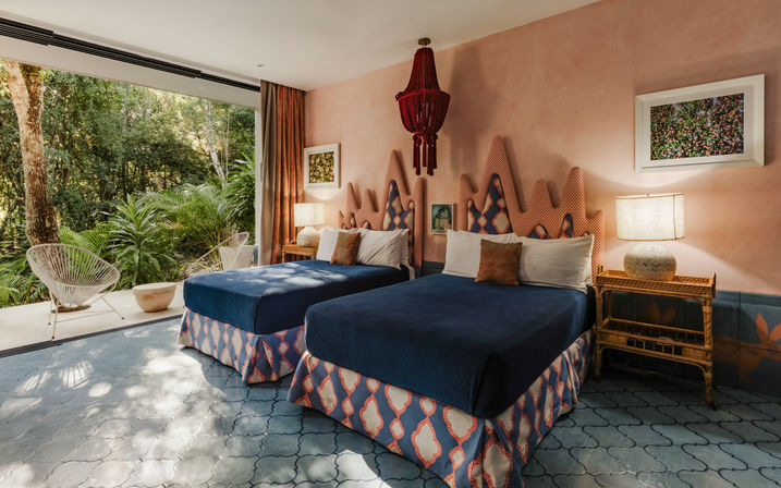 Boho-chic tropical retreat bedroom with two navy twin beds, zigzag patterned headboards, rattan nightstand and lamps, red pendant light and a sliding wall opening to a lush forest patio with white wire chairs