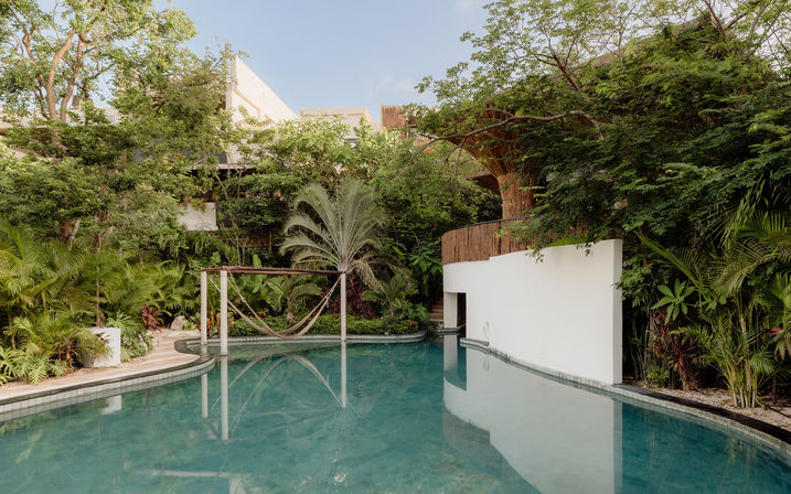 Secluded tropical pool oasis with calm turquoise water, a hammock strung between wooden posts, dense palm and jungle foliage, and a curved white modern villa wall.