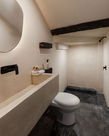Minimalist rustic bathroom with long concrete vanity and integrated sink, round wall mirror, black wall-mounted faucet, white toilet, dark stone tile floor and exposed wooden beams.