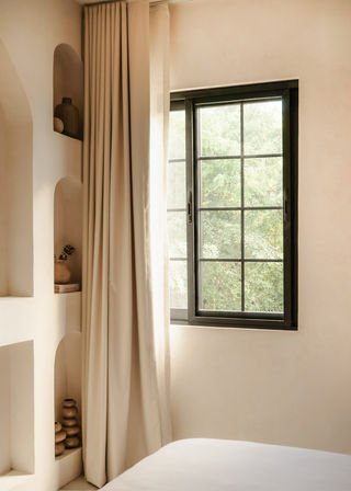 Sunlit bedroom corner with floor-length beige curtains, black grid-framed window overlooking green trees, and recessed plaster niches displaying ceramic vases.