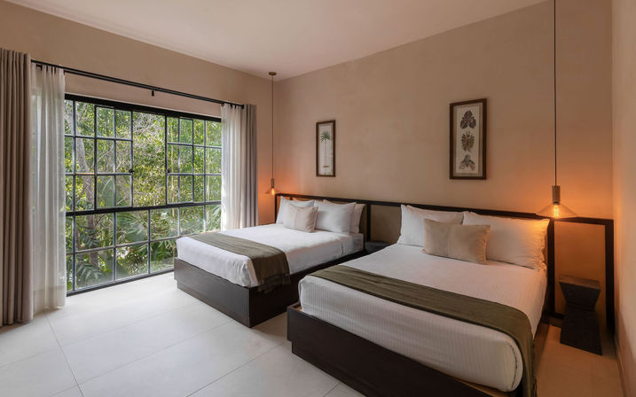 Modern hotel room with two beds, neutral beige walls, olive-green bed runners, large black-framed window opening to lush tropical greenery, sheer curtains, hanging amber pendant lights and framed botanical prints.