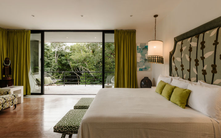 Modern bedroom with king bed, patterned green headboard and pillows, sliding glass doors to a balcony overlooking a lush tropical forest, green curtains and pendant lamp.
