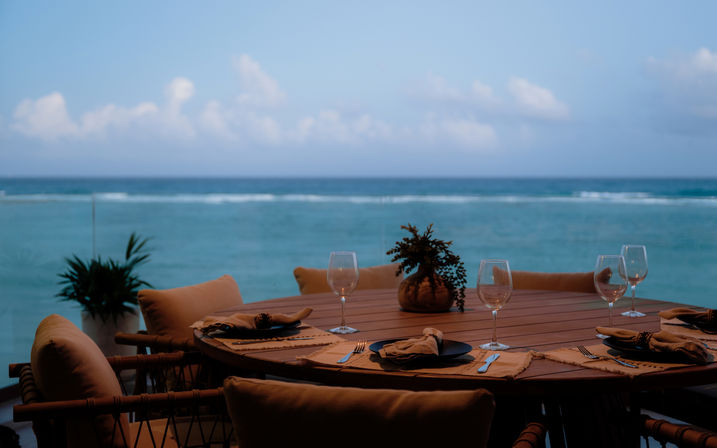 Cozy oceanfront dining table set for four on a wooden seaside terrace with wine glasses, napkins and a small centerpiece, turquoise ocean and cloudy sky in the background.