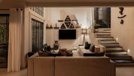 Contemporary neutral living room interior — cozy sectional sofa facing a wall-mounted flat-screen TV with triangular shelves, decorative vases, large grid windows, ambient floor lamp and a floating staircase.