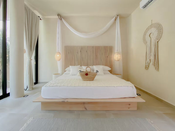Sunlit minimalist coastal bedroom with wooden platform king bed, sheer canopy, hanging rattan pendants, woven wall art, straw basket on crisp white linens and floor-to-ceiling glass doors with light curtains.