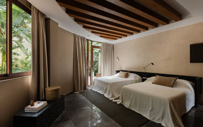 Sunlit tropical retreat with twin beds and white linens, exposed wooden beam ceiling, floor-to-ceiling windows opening to lush greenery and neutral minimalist decor.