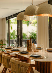 Sunlit indoor-outdoor dining room with a long wooden table set for six—woven rattan chairs, black plates on straw placemats, stemmed glasses, rustic ceramic vases, glowing textured dome pendant lights, and glass doors opening to a lush palm garden.