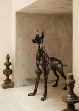 Stoic bronze Great Dane sculpture standing in a neutral plaster niche with antique finials nearby — minimalist interior decor accent for a modern foyer.
