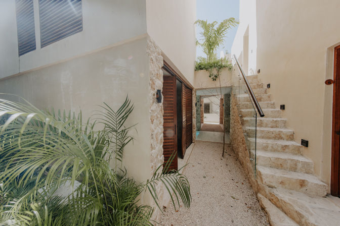 Sunlit tropical courtyard passage with gravel walkway, stone staircase and glass railing, wooden louvered doors and lush palm plants.