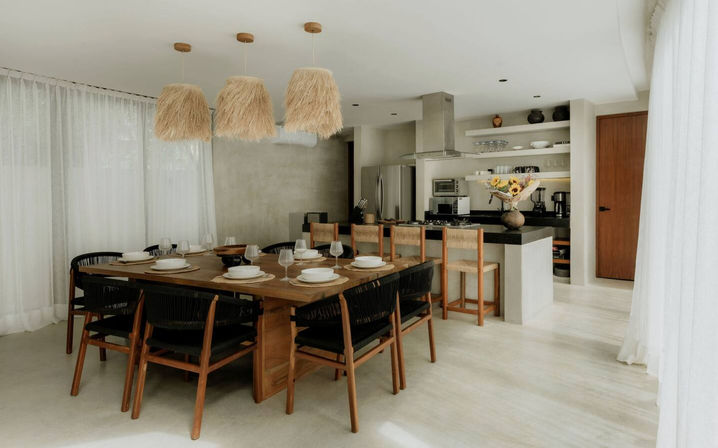 Open-plan modern dining room and kitchen with large wooden table, black woven chairs, three rattan pendant lights, kitchen island with bar stools and neutral coastal-boho decor.