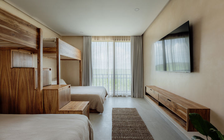 Sunlit modern hotel room with natural wood bunk and platform beds, large wall-mounted TV, sheer curtains over sliding glass doors opening to a green balcony view, neutral linens and woven rug.